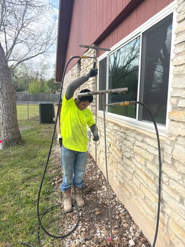 A technician guiding two soil injection rods into the ground beside a stone clad foundation, with a hose feeding each rod.